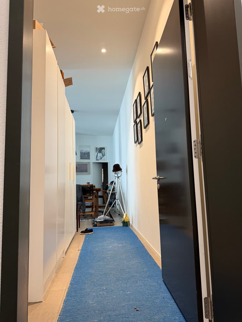 A narrow hallway with light-colored walls, a blue runner carpet, and several framed pictures on the walls. A door is open to reveal a room with a table, chairs, and a tripod.