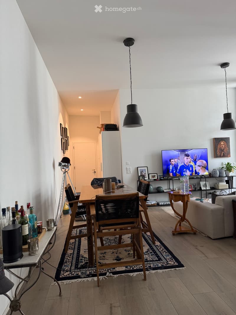 A modern living and dining area with a sofa, dining table, chairs, television, pendant lights, shelves, and decorative items.