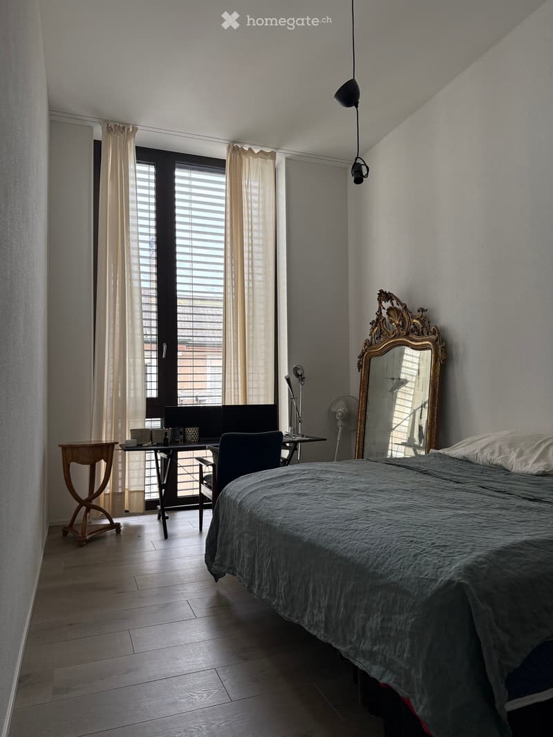 Modern bedroom with a wooden floor, large window with blinds and curtains, a bed with a grey duvet, a mirror with an ornate frame, a small table with a laptop, a chair, and a fan.