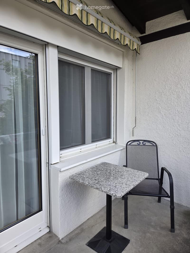 Balcony with a granite table and a black metal chair, large windows with white frames, striped awning above.