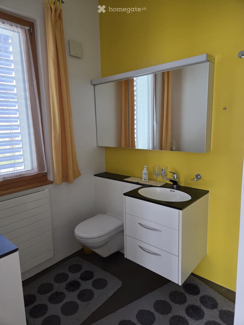 Bright bathroom with yellow walls, white cabinetry, a sink, toilet, and a large mirror above the sink. A window with blinds and a curtain is on the left side.