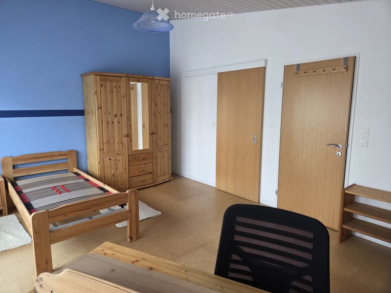 A furnished bedroom with a wooden bed, wardrobe, and a black chair. The room features light wooden flooring and white walls with blue accents.