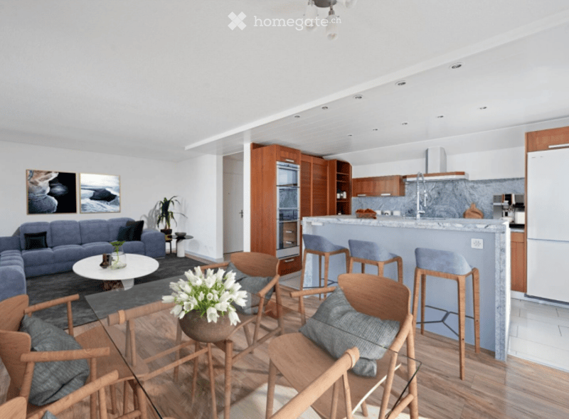 Open-plan living and dining area with a modern kitchen. Features a grey sofa, coffee table, bar stools, and a kitchen island with bar stools. The space includes a potted plant and picture frames on the wall.