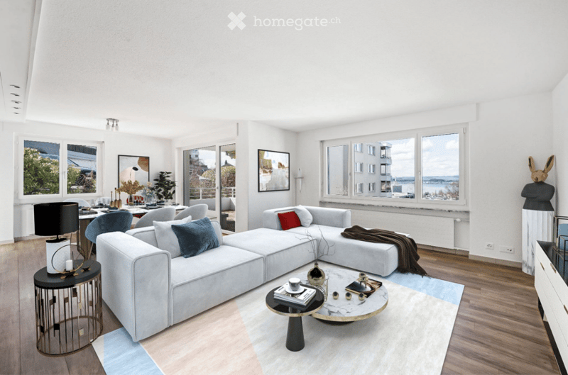 Modern living room with large windows offering a view, featuring a grey sectional sofa, coffee table, side table with lamp, dining area with chairs and table, and decorative items including a picture frame and rug.