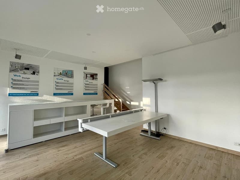 Modern reception area with a white reception desk, two chairs, a table, and shelves on the wall. The floor is wooden, and the walls are white with posters.