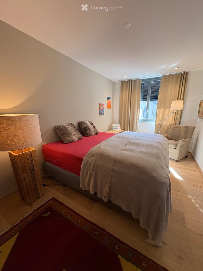 Cozy bedroom with a double bed, red and grey bedding, wooden floor, side table with lamp, armchair, and window with curtains.