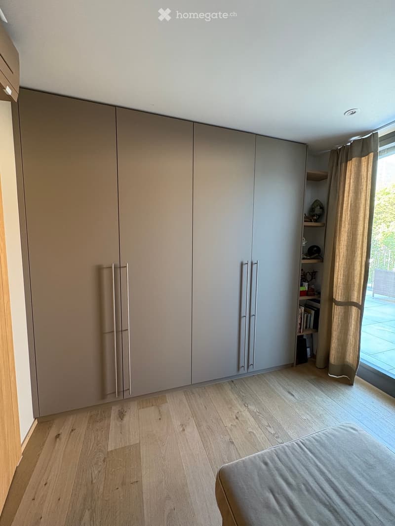Modern bedroom with light wood flooring, large built-in wardrobe with silver handles, and a small shelf unit near the window with books and decorative items.