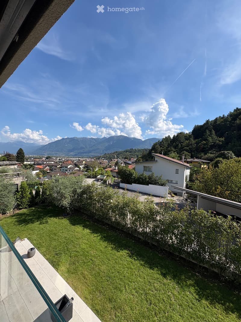 Garden with green grass, surrounded by bushes and trees, overlooking a town with mountains in the background.