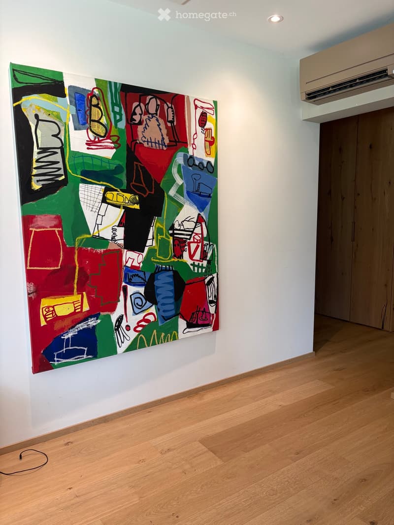 Empty room with light wooden flooring and white walls, featuring a large abstract painting on one wall and an air conditioning unit on the adjacent wall.