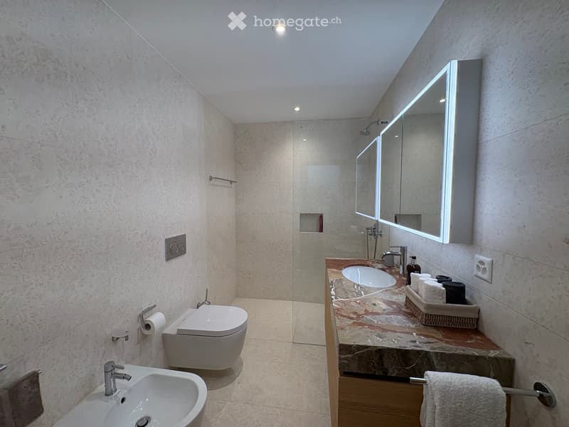 Modern bathroom with marble walls and floors, featuring a double sink vanity, a toilet, a large illuminated mirror, and neatly arranged towels.