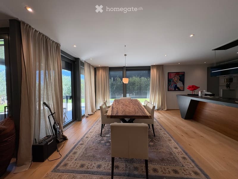Modern dining area with a wooden dining table, white chairs, a large window with curtains, a kitchen counter, and a guitar leaning against the wall.