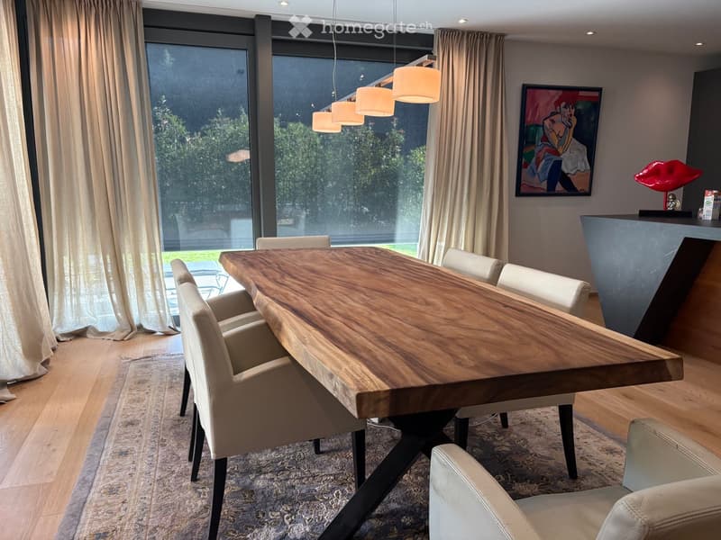 Modern dining room with a large wooden dining table, white chairs, large windows with curtains, pendant lights, and a painting on the wall.