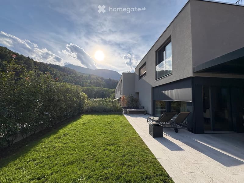 Modern house with a well-maintained lawn, patio area, lounge chairs, and a scenic mountain view in the background.
