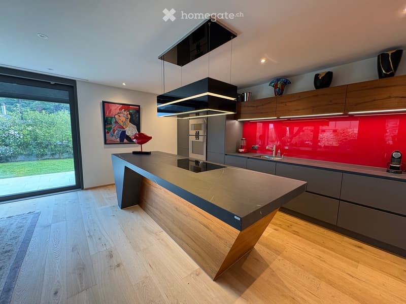 Modern kitchen with a large island, stainless steel appliances, and a red backsplash. Features pendant lights, a large window, and decorative artwork.