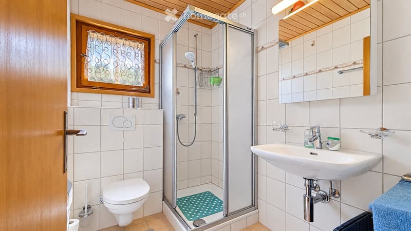 Modern bathroom with white tiles, a toilet, a sink with a mirror, a glass-enclosed shower, and a window with a curtain.