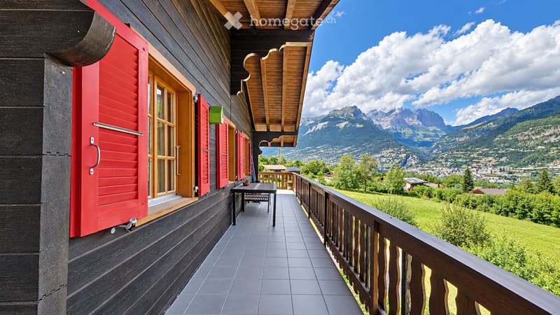 Balcony with wooden railing, tiled floor, and a view of mountains and a town below. Features a small table and chair.