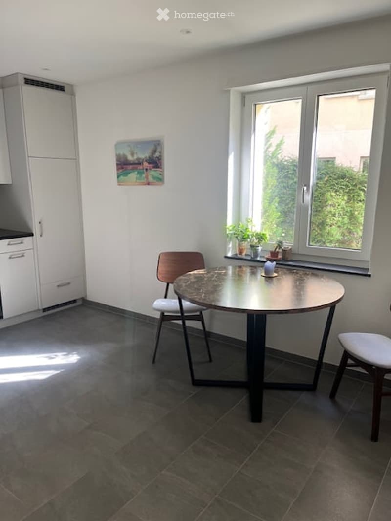 Bright room with a round table, two chairs, white cabinets, and potted plants on the windowsill. A picture frame hangs on the wall.