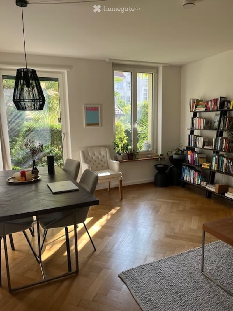 Bright, modern dining area with wooden flooring, large windows, a dark wooden dining table, chairs, a white armchair, a bookshelf, and a floor lamp.