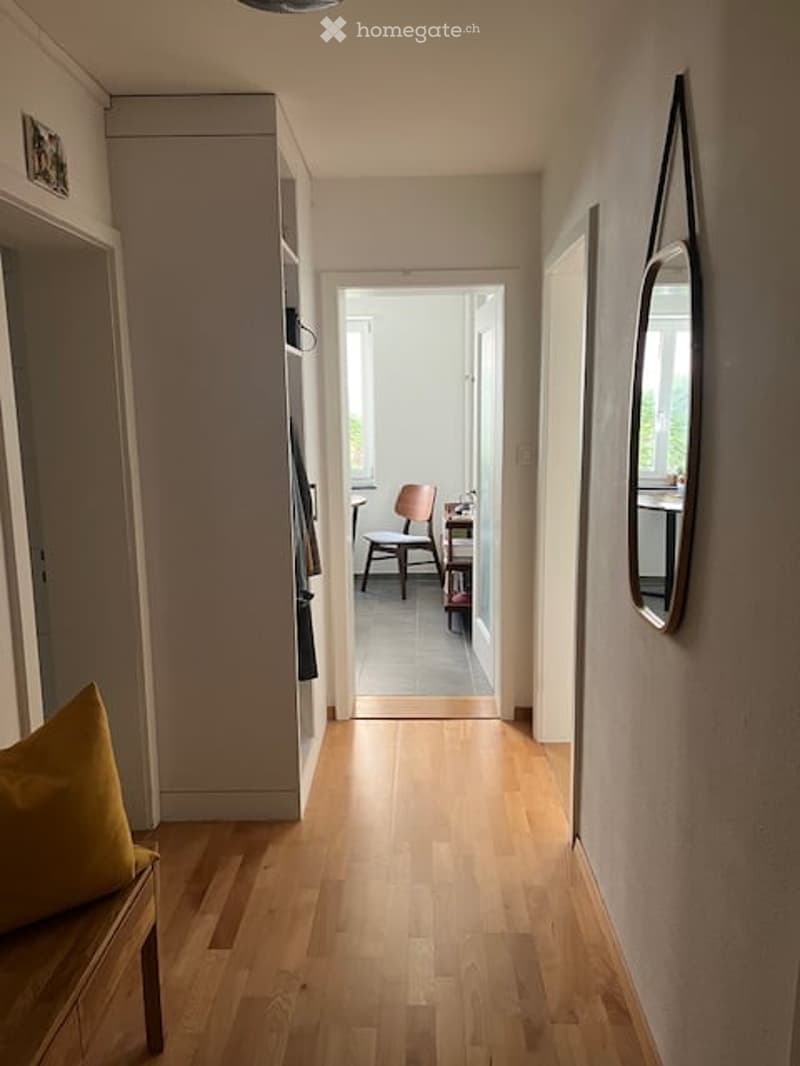 A narrow hallway with light wooden flooring, white walls, and a bench with a yellow cushion on the left side. A mirror is mounted on the right wall, and at the end of the hallway, a small room is visible with a chair and a table.