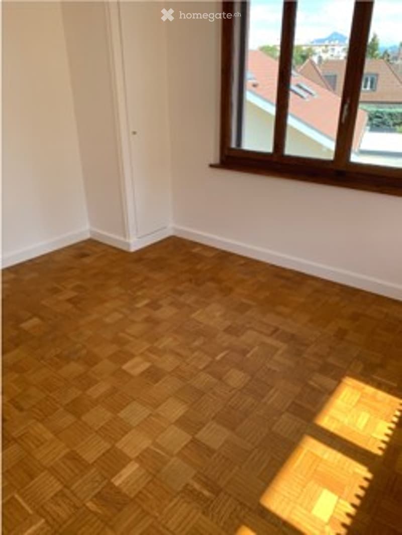 Empty room with wooden herringbone flooring, white walls, and large window with view of rooftops.