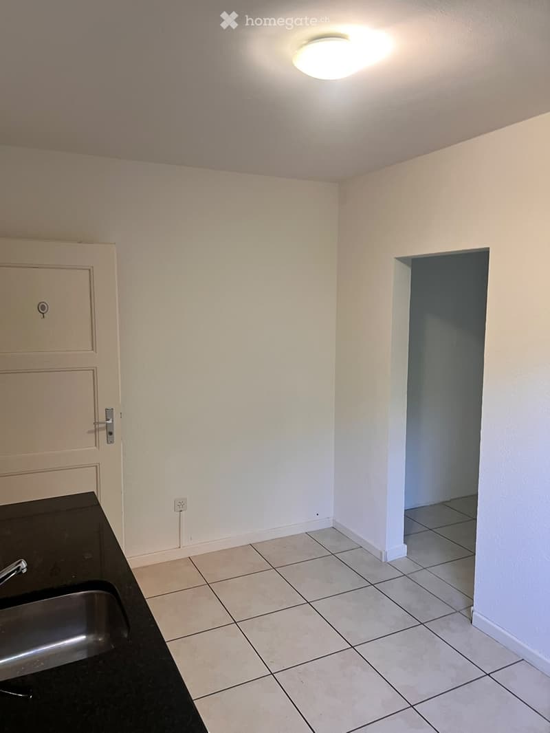 Empty room with tiled floor, white walls, a door, and a ceiling light fixture. A small black countertop with a sink is visible on the left side.