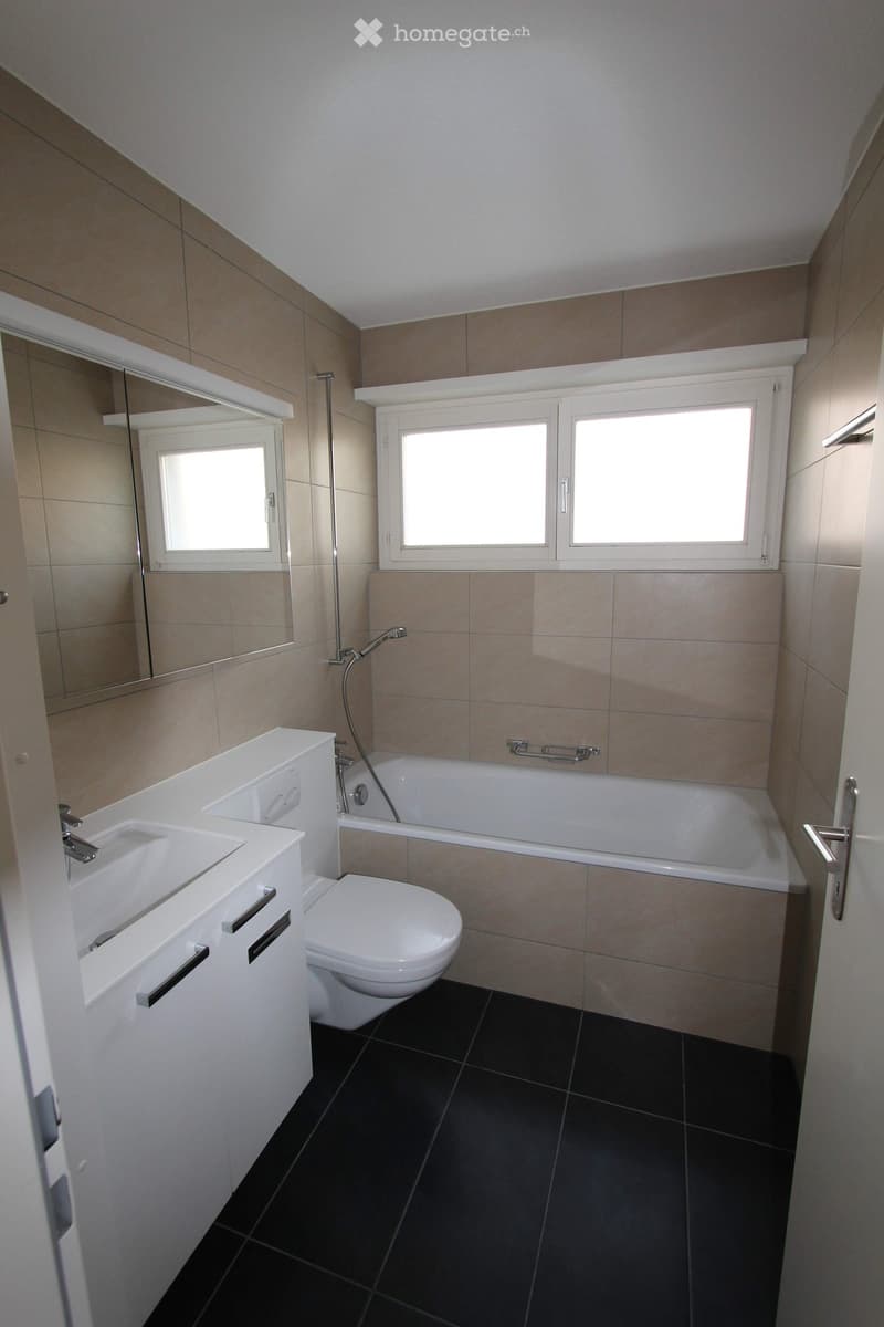 Modern bathroom with a white sink, toilet, bathtub, and large mirror. Features dark tiled flooring and beige tiled walls.