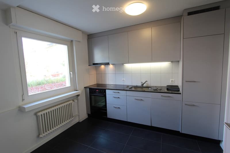Modern kitchen with white cabinets, black countertop, sink, oven, and a window overlooking a garden area.