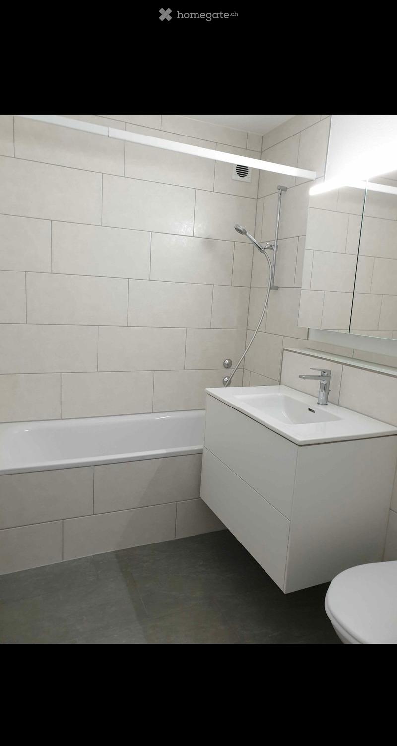 Modern bathroom with a bathtub, sink, toilet, and showerhead. Features light-colored tiles and a sleek design.