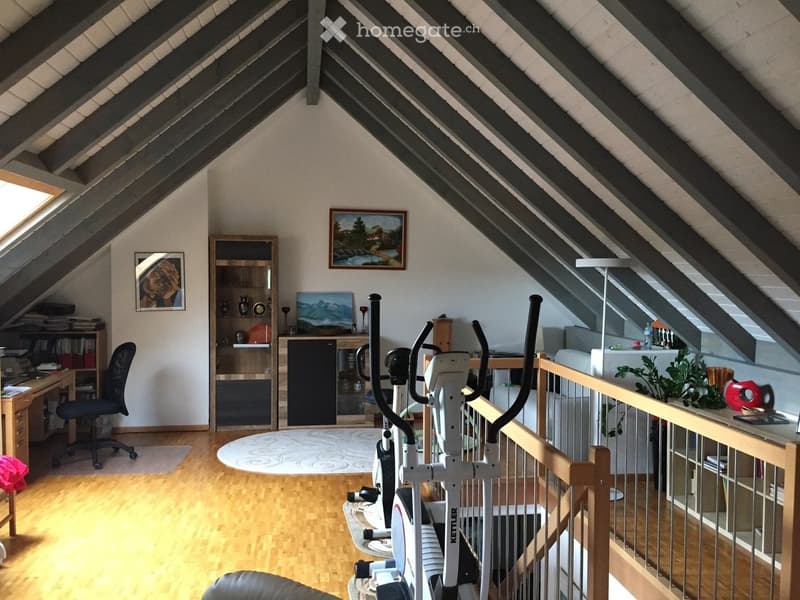 Spacious attic room with wooden flooring, slanted ceilings, and large windows. Features a desk with a chair, bookshelf, exercise bike, and a small seating area with a stool and painting on the wall.