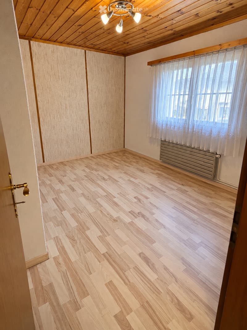 Empty room with wooden flooring, light-colored walls, and a window with white curtains. The room features a radiator below the window and a wooden ceiling with a light fixture.