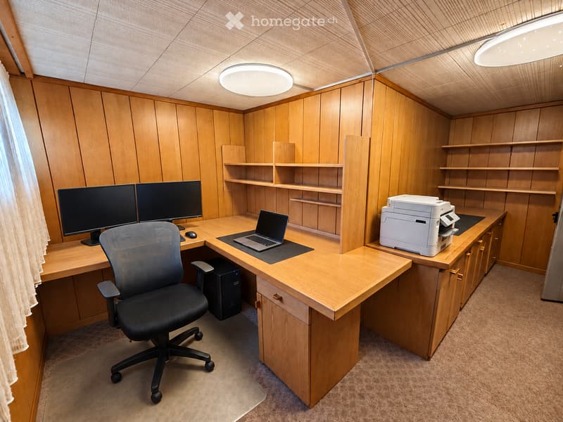 A modern office space with wooden walls and ceiling, featuring a large desk with a laptop, two computer monitors, a printer, and a comfortable office chair.