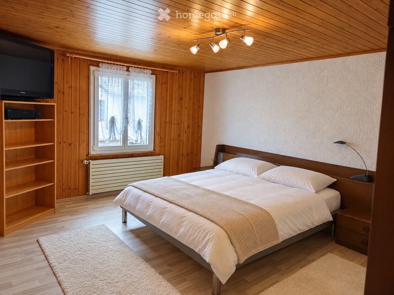 Cozy bedroom with wooden walls and ceiling, featuring a large bed with white bedding, a nightstand with a lamp, a shelf with a TV, and a radiator beneath a window with sheer curtains.