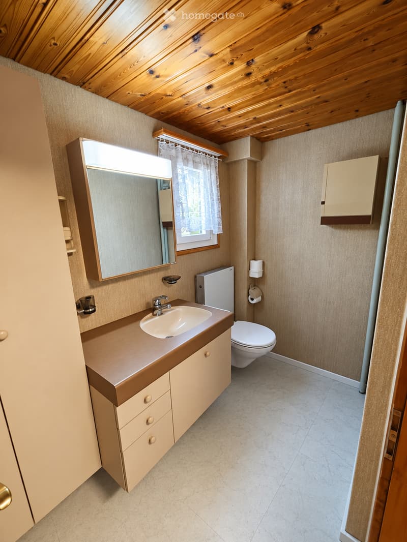 Bathroom with wooden ceiling, beige walls, sink with faucet, toilet, mirror, and cabinet.
