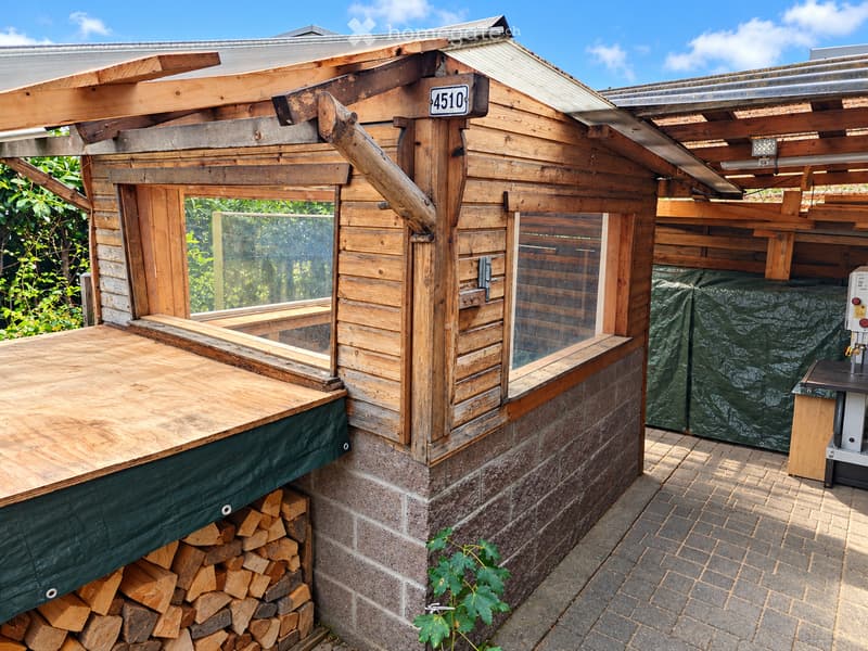 Wooden outdoor structure with a covered deck, glass windows, and a stone base. Includes a stack of firewood and a covered grill.