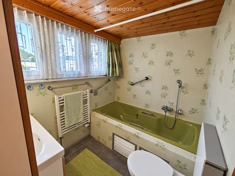 Bathroom with green bathtub, white toilet, towel rack, sink, and window with curtains. Features wooden ceiling and floral tile design.