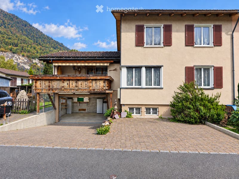 Two-story house with beige exterior, red shutters, and a wooden balcony. Features a garage and is situated in a scenic, mountainous area.