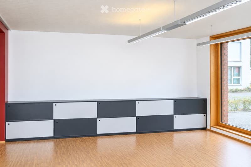 Modern, minimalist room with light wooden flooring, white walls, and a large grey and white cabinet unit against one wall. The room features recessed ceiling lighting and large windows with wooden frames, offering a view of the outside.