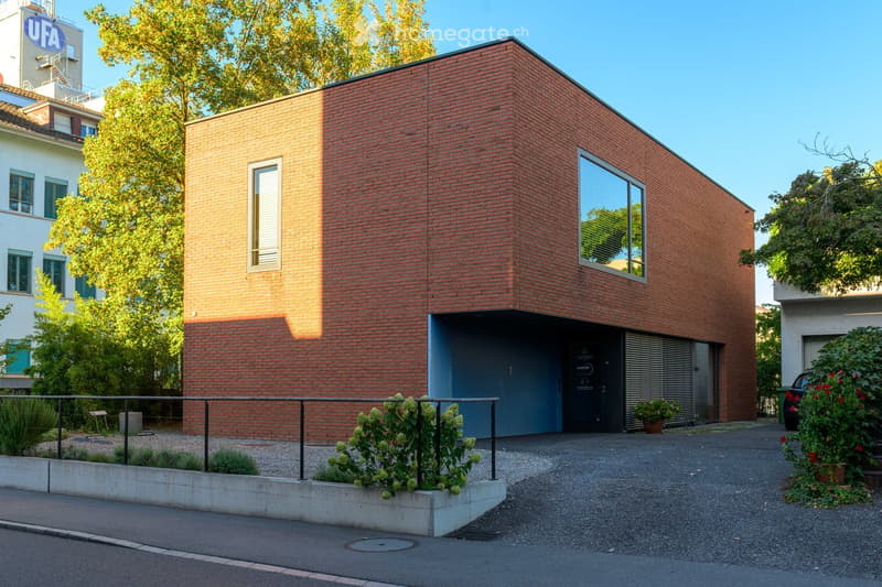 Modern red brick building with a flat roof, large windows, and a blue garage entrance. The building is surrounded by greenery and a paved driveway.