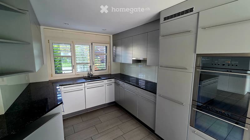 Modern kitchen with white and grey cabinets, black countertops, and a stainless steel oven. Features a sink with a faucet and large windows with blinds.
