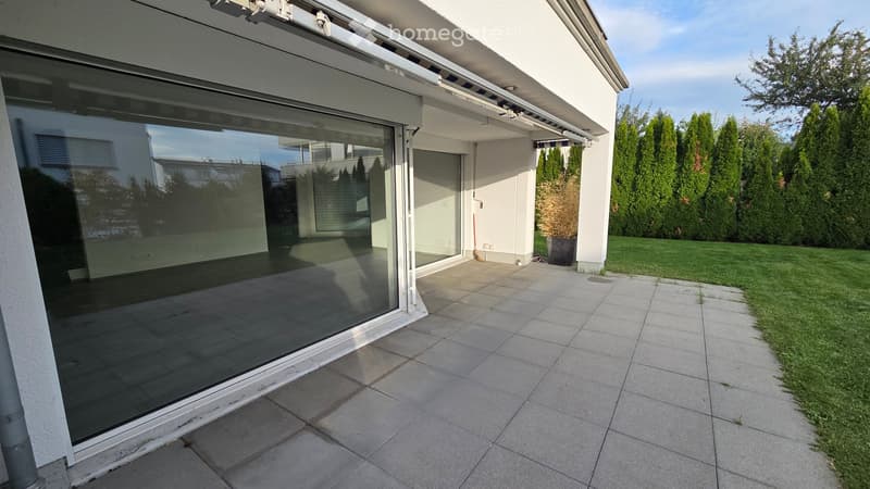 Modern glass sliding door leading to a tiled balcony area with a view of the garden and surrounding greenery.