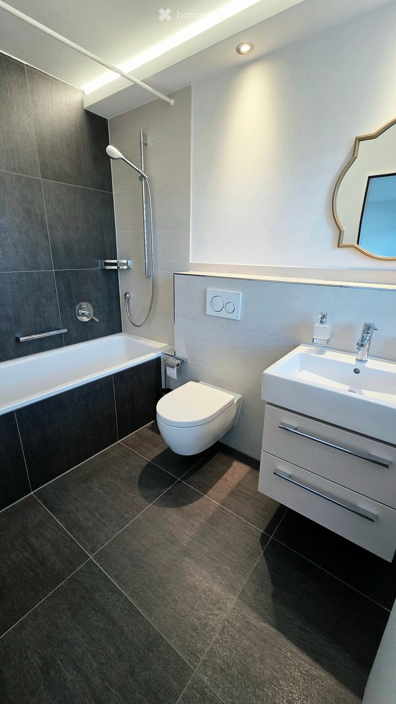 Modern bathroom with dark grey tiles, white fixtures, a bathtub, toilet, sink, and a large mirror with a decorative frame.