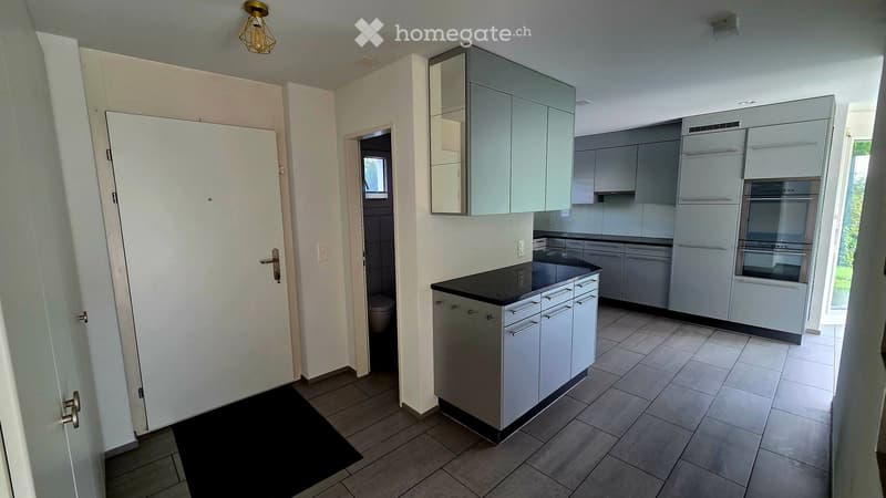 Modern kitchen with grey cabinets, black countertop, stainless steel appliances, and a visible bathroom area through an open doorway.