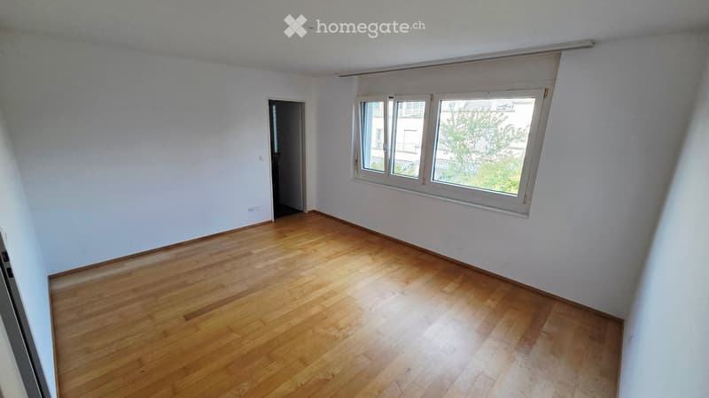 Empty room with wooden flooring, white walls, and large windows with blinds. There is a doorway leading to another room.