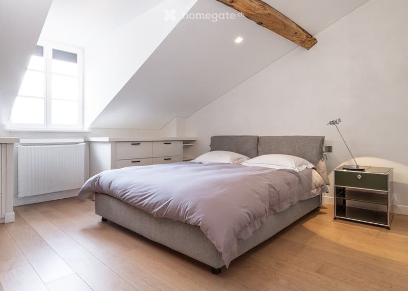 Bright, modern bedroom with wooden flooring, a large bed with grey bedding, a nightstand with a lamp, and built-in cabinets.