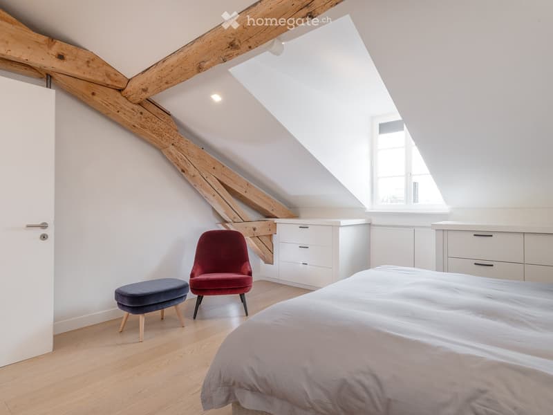 Bright attic bedroom with wooden beams, white walls, and large windows. Features a bed, red velvet chair, blue ottoman, and white built-in cabinets.