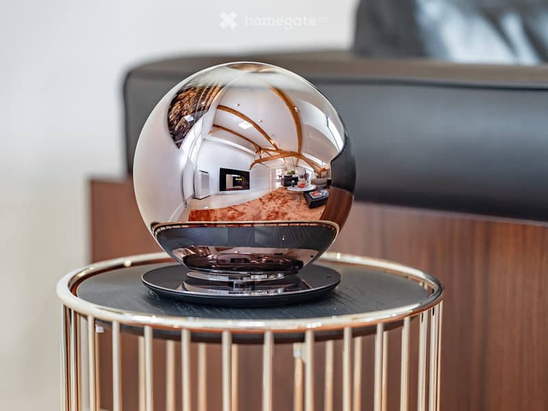 A reflective globe sits on a round table in a modern living room, with a sofa, coffee table, and television visible in the background.