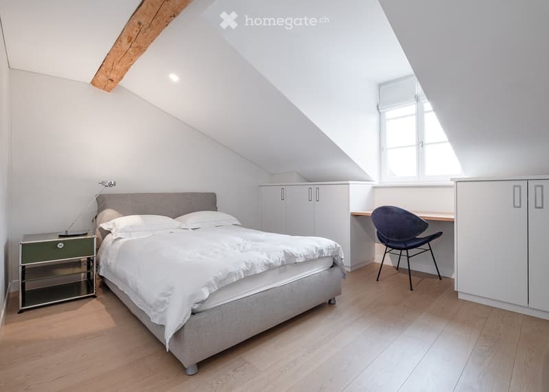 Bright, modern bedroom with a large bed, nightstand, desk, chair, and built-in cabinets. The room features wooden flooring and large windows allowing natural light to fill the space.
