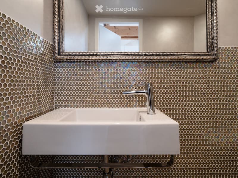 Bathroom with a white sink, chrome faucet, and a large mirror with a decorative frame. The walls are covered in mosaic tiles.