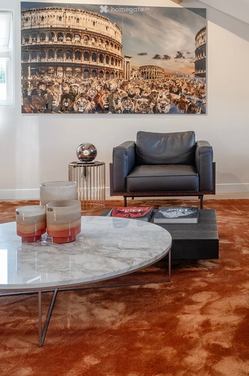 Modern living room with a large wall art depicting the Colosseum, a sleek black leather armchair, a round glass coffee table with decorative candle holders, and a small side table with a lamp and books.