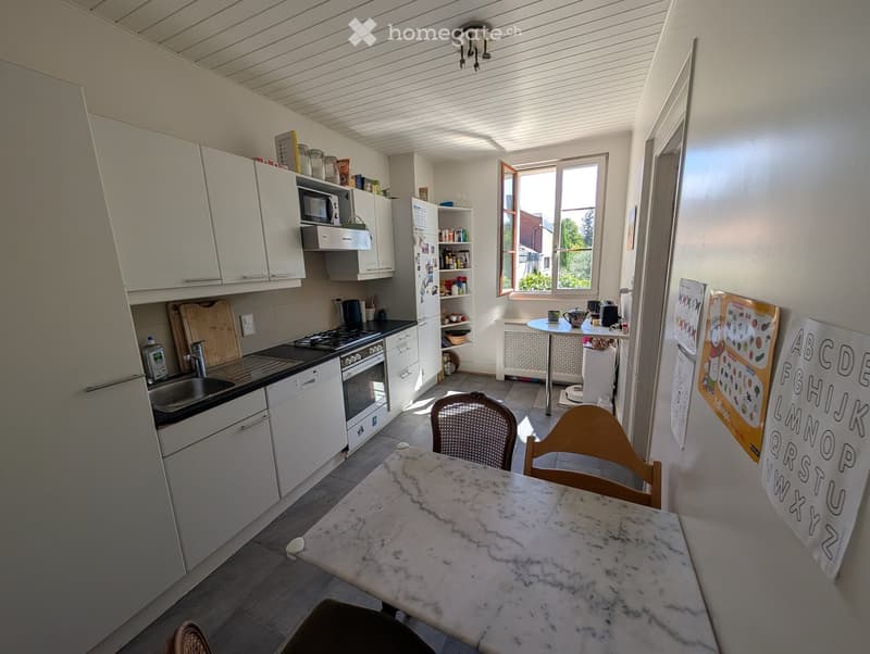 Bright kitchen with white cabinets, stainless steel appliances, marble dining table, and two chairs. Features a window with a view of greenery.